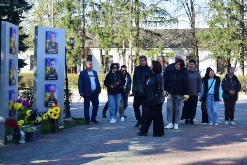 Група людей на вулиці проходить повз пам'ятні стели з портретами військових. Біля підніжжя меморіалу лежать квіти. На фоні видно дерева та будівлю, а подія відбувається в сонячний день.