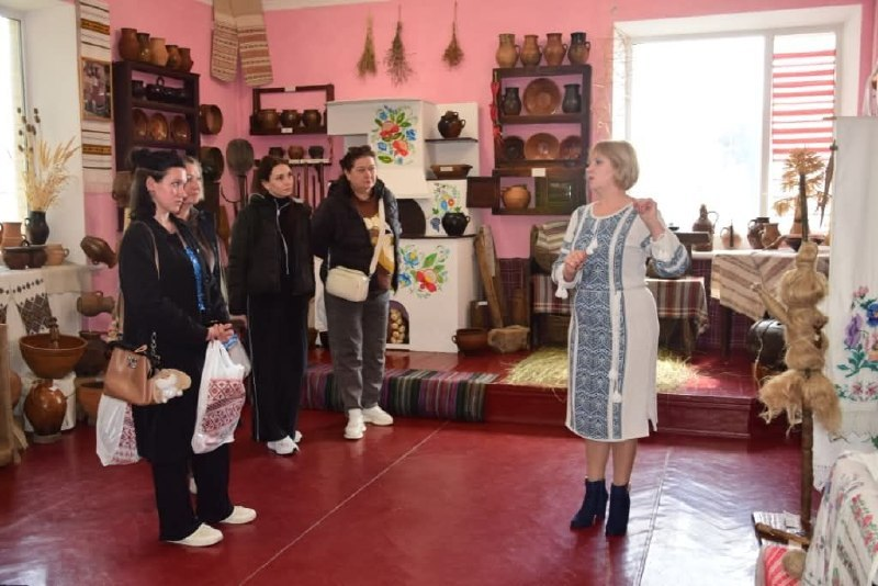 Жінка у вишиванці проводить екскурсію в музеї народного побуту. В інтер'єрі представлені традиційна піч, керамічний посуд та вишиті рушники.