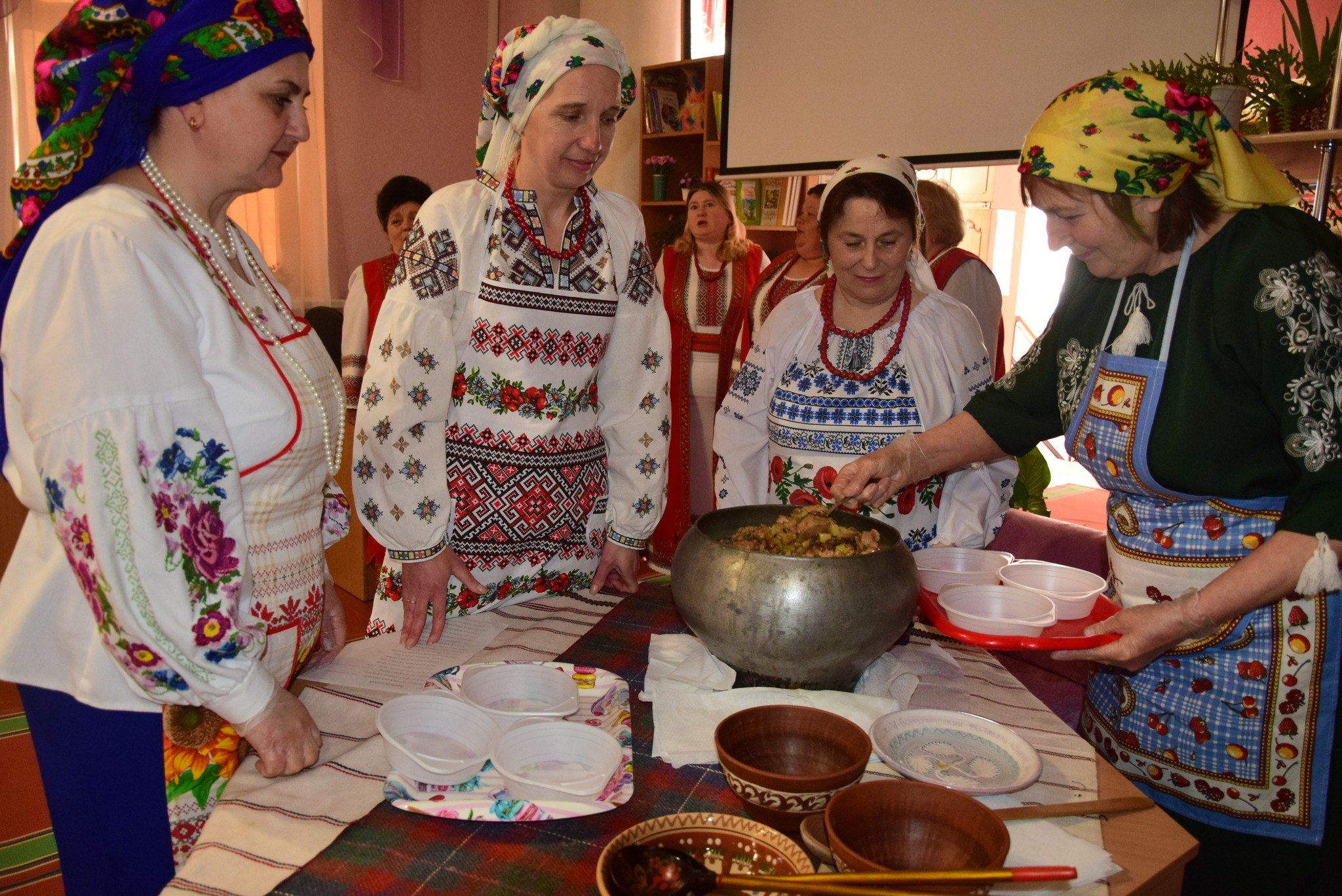 Жінки в українському національному вбранні та хустках зібралися біля столу. Одна з них насипає страву з великого чавунного казана в порційні миски. На столі лежать вишиті рушники та дерев'яний посуд.