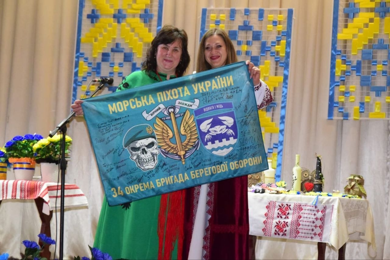 Дві жінки на сцені тримають прапор Морської піхоти України з підписами та військовою символікою. Тло прикрашене декораціями у національних кольорах, а поруч стоїть стіл із вишитою скатертиною та пам'ятними речами.