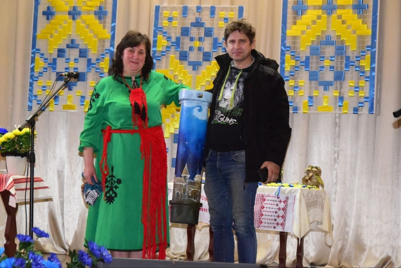 Жінка у вишитій сукні та чоловік на сцені стоять поруч із високим розмальованим предметом, схожим на гільзу. Тло прикрашене синьо-жовтими декораціями та вишитими рушниками.