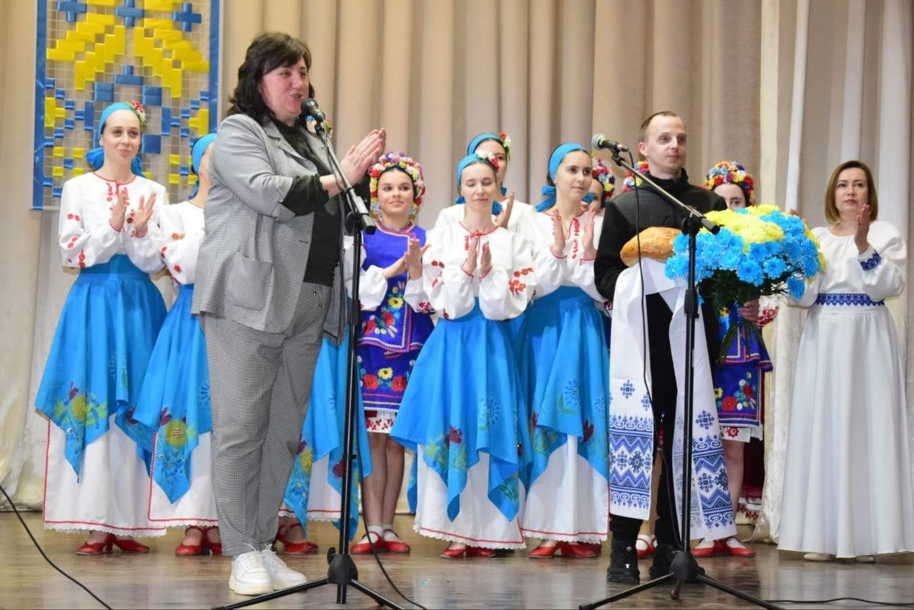 Жінка виступає біля мікрофона на сцені перед групою молоді в українських народних костюмах. Поруч стоїть чоловік із короваєм на вишитому рушнику та великим синьо-жовтим букетом квітів.