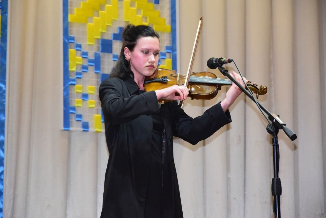 Дівчина в чорному вбранні грає на скрипці біля мікрофона на сцені. Тло прикрашене світлою завісою та декорацією в національних кольорах.