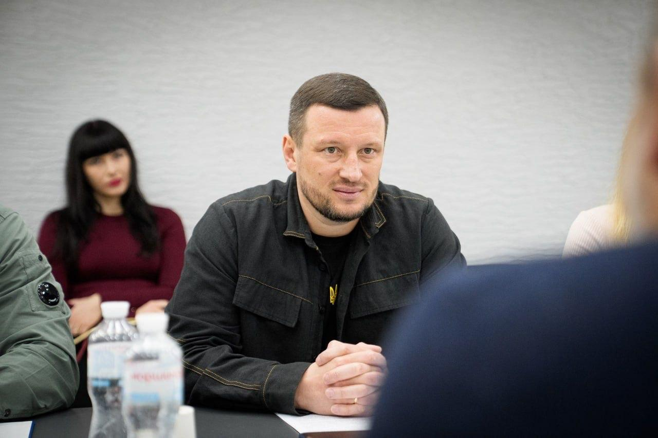 Чоловік у темній сорочці сидить за столом, склавши руки перед собою, та уважно слухає співрозмовника під час зустрічі. На задньому плані видно жінку та робочу обстановку.