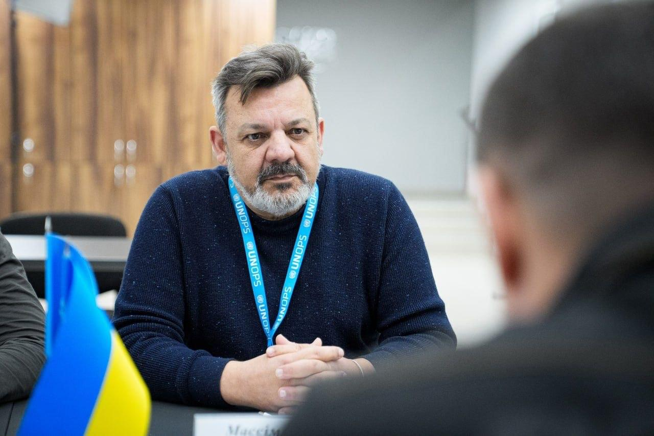 Чоловік із сивою бородою в синьому светрі та з бейджем на шиї сидить за столом під час розмови. На передньому плані видно невеликий прапор України та частину фігури іншої людини.