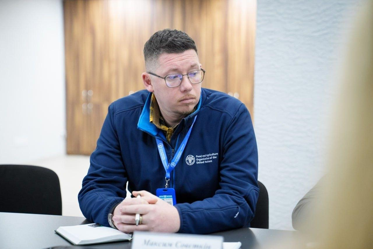 Чоловік в окулярах та синій куртці з емблемою міжнародної організації сидить за столом. Перед ним лежить розгорнутий блокнот, він тримає ручку та уважно дивиться вбік під час обговорення.