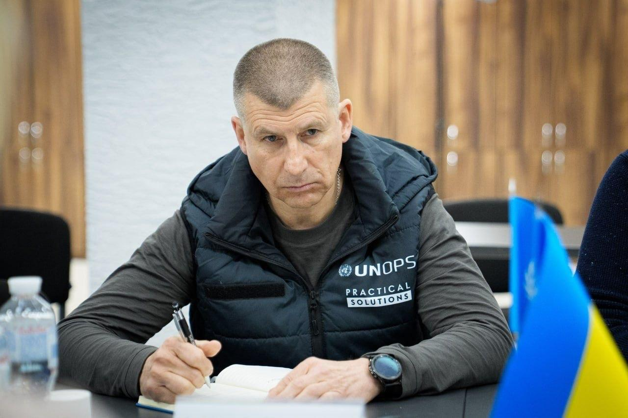 Чоловік у чорному жилеті з логотипом UNOPS робить записи в блокноті під час зустрічі. Він зосереджено дивиться перед собою; на столі поруч видно прапор України та пляшку води.