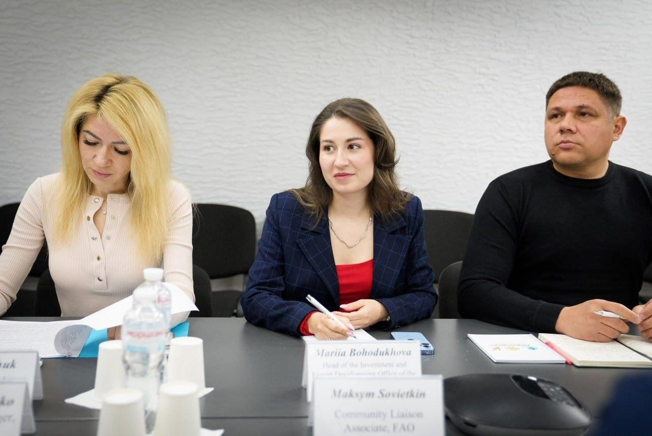 Три людини за столом під час наради. Жінка в центрі посміхається, учасники тримають ручки та працюють із документами. На передньому плані — таблички з іменами та пляшки з водою.