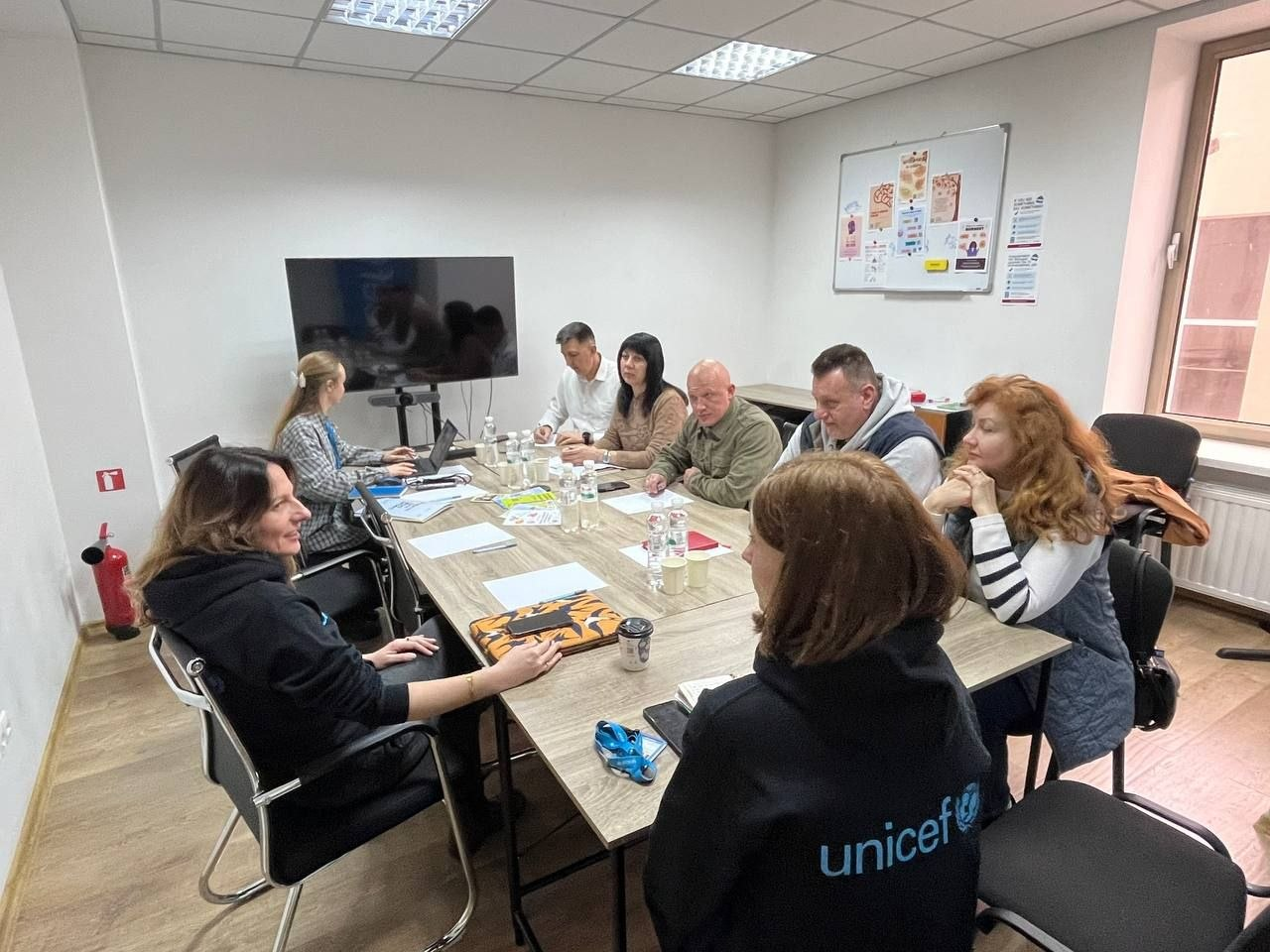 Група людей за великим столом під час робочої наради в офісному приміщенні. У кімнаті є телевізор, маркерна дошка з плакатами та вікно. На передньому плані — людина в чорному худі з логотипом UNICEF.