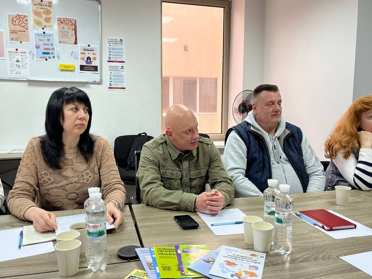 Група людей за робочим столом під час обговорення в офісному приміщенні. На столі — пляшки з водою, паперові стаканчики та друковані інформаційні матеріали. На стіні позаду розміщена дошка з плакатами.