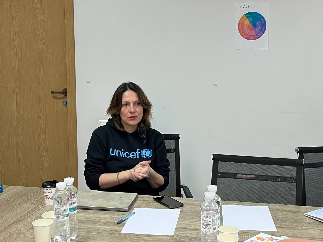 Жінка в чорному худі з логотипом UNICEF за столом під час робочої зустрічі. На столі перед нею — документи, пляшки з водою та ноутбук. На стіні позаду розміщено кольорову інфографіку у формі кола.