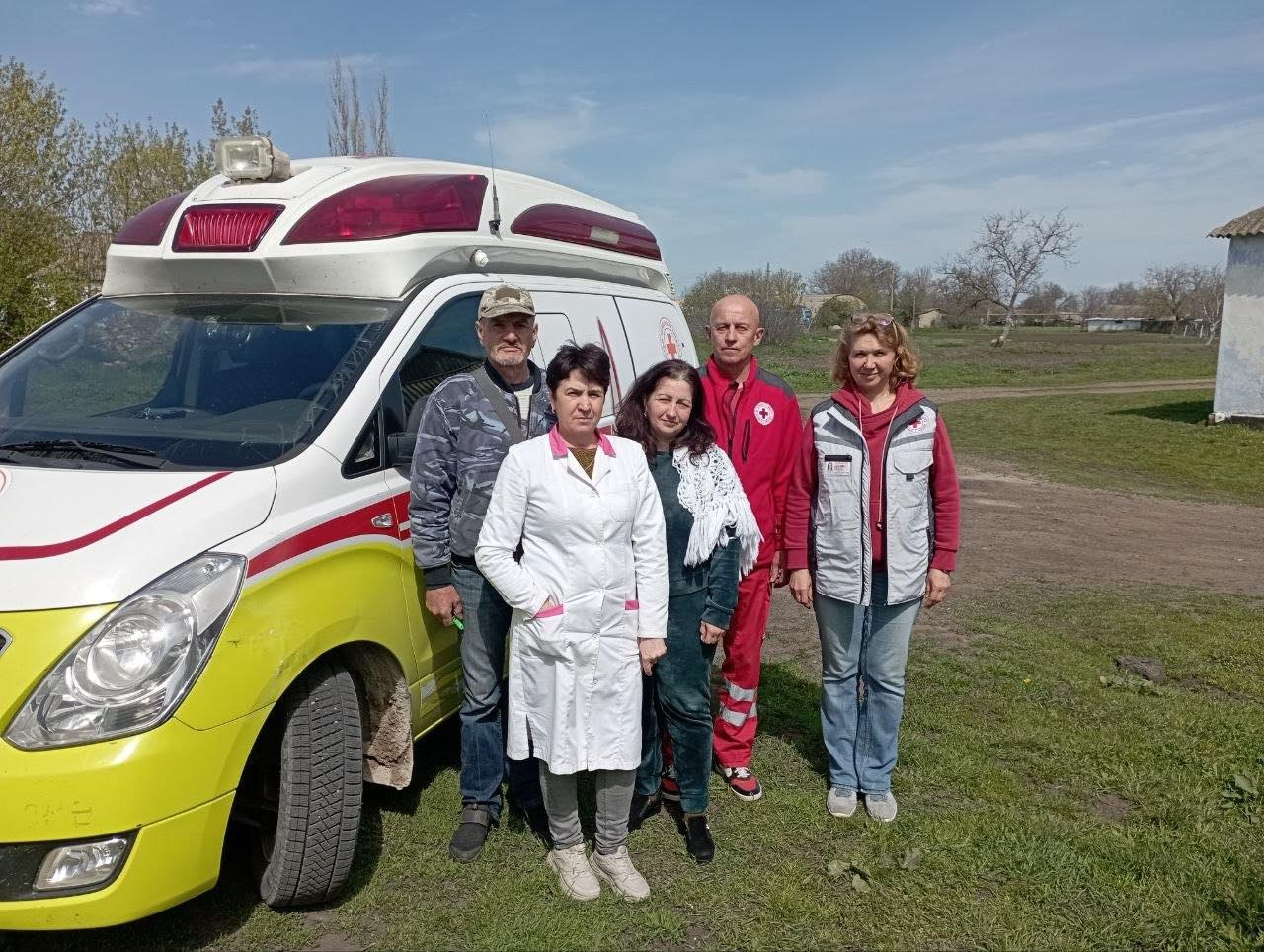 Група медичних працівників та волонтерів, серед яких представники Червоного Хреста, стоять поруч із медичним автомобілем на відкритій місцевості.
