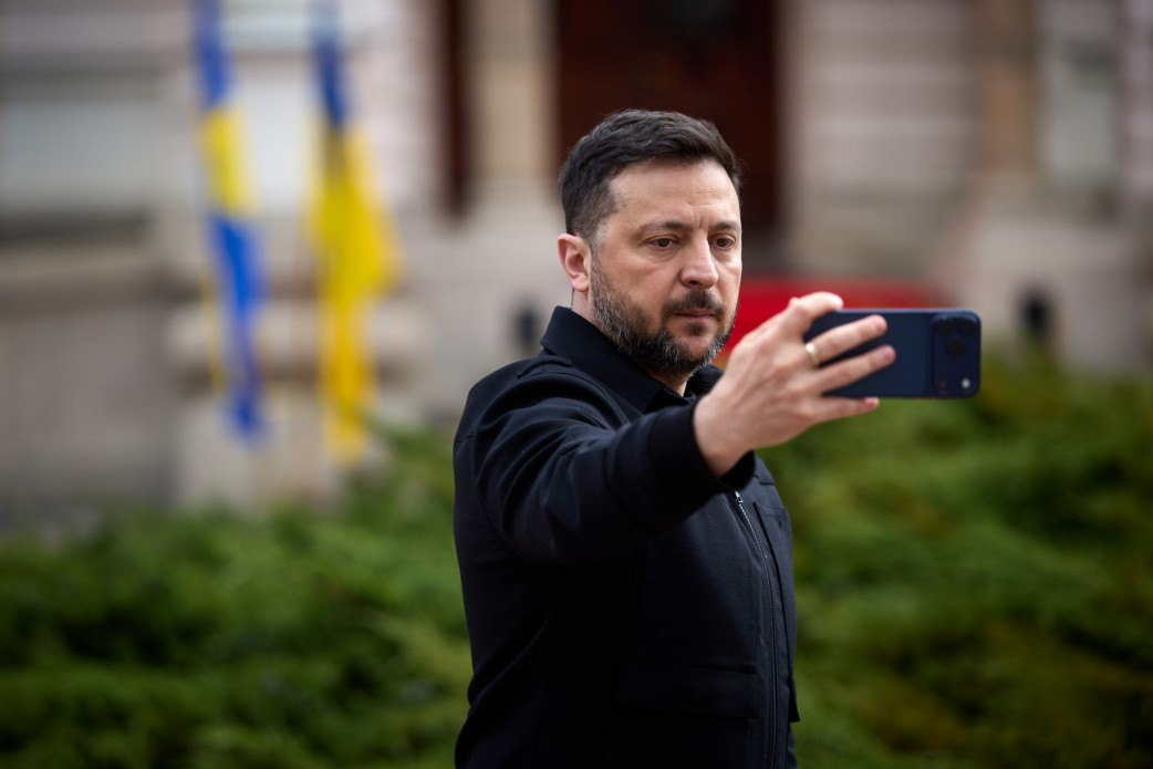 Чоловік у чорному одязі знімає відеоселфі на смартфон на фоні будівлі та прапорів.