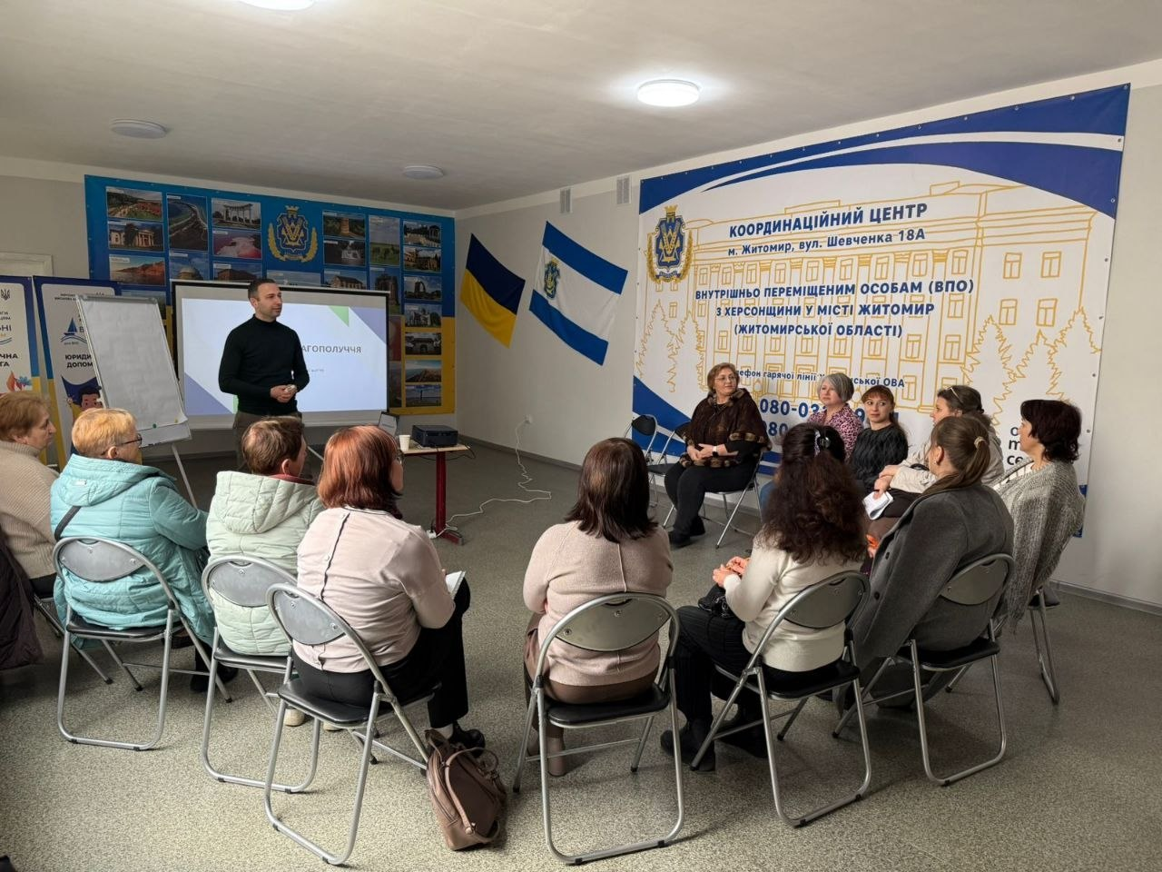 Група людей на інформаційній зустрічі в офісному приміщенні. Чоловік стоїть біля екрана презентації та фліпчарта, звертаючись до аудиторії, що сидить півколом. На стіні розміщено великий інформаційний банер та прапори.