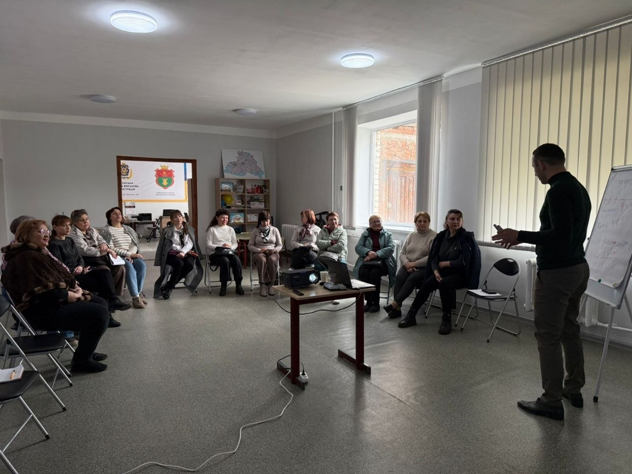 Чоловік проводить презентацію для групи людей, які сидять півколом у світлій залі. У центрі стоїть проєктор на столику, а поруч — фліпчарт із записами.