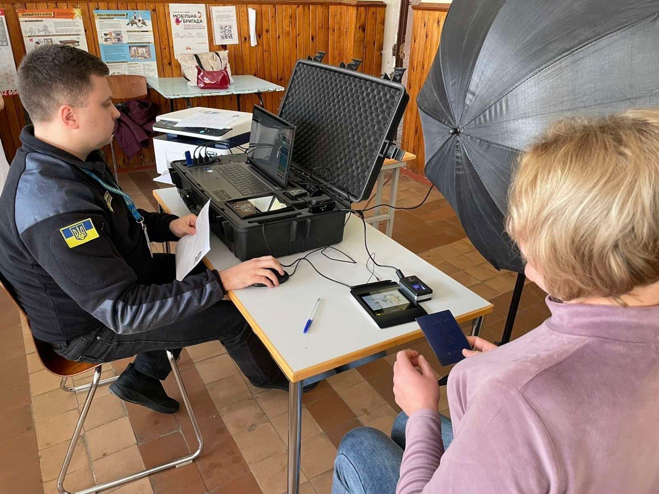 Працівник у формі з українською символікою надає послуги жінці за допомогою мобільного кейса з технікою. На столі розміщено ноутбук у захисній валізі, сканер відбитків пальців та інше обладнання, а поруч встановлено фотопарасолю.