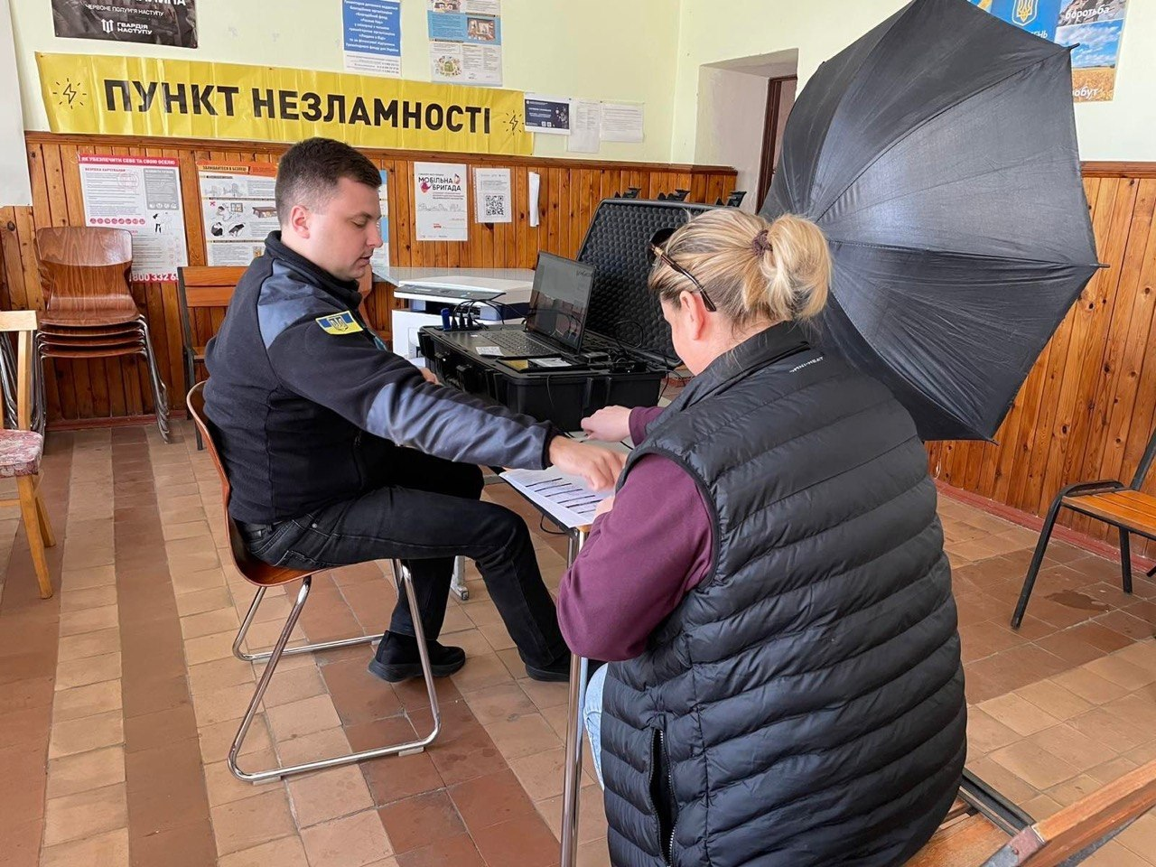 Працівник у формі з українською символікою за допомогою мобільного кейса з обладнанням надає послуги жінці в приміщенні «Пункту незламності». На столі стоїть ноутбук у захисній валізі, а поруч встановлено фотопарасолю.