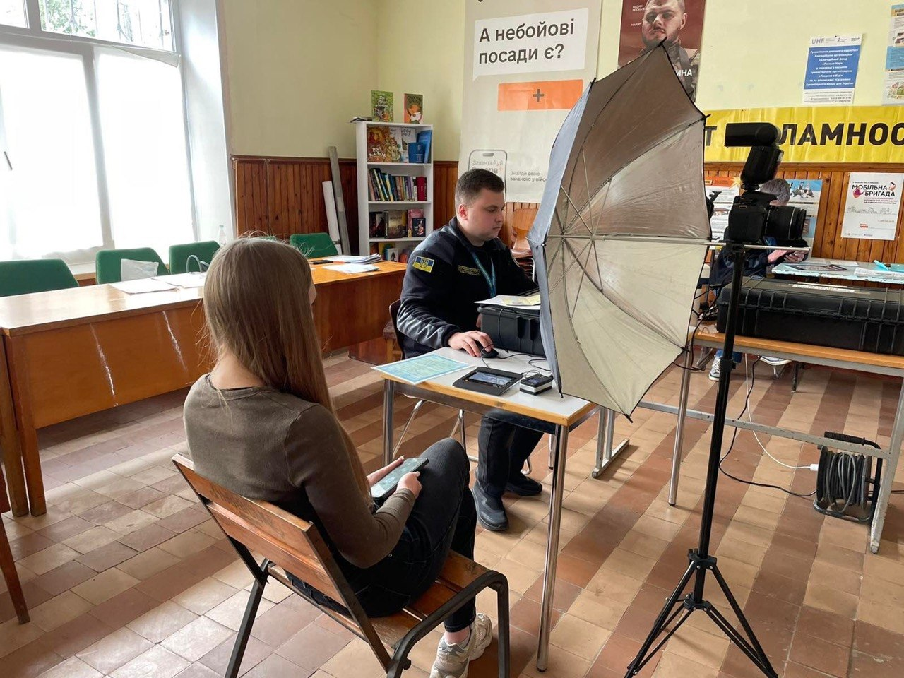 Процес оформлення документів у приміщенні «Пункту незламності». Працівник у формі працює за столом із мобільним кейсом та обладнанням, навпроти нього сидить відвідувачка. Поруч встановлено стійку з фотопарасолею та камерою.