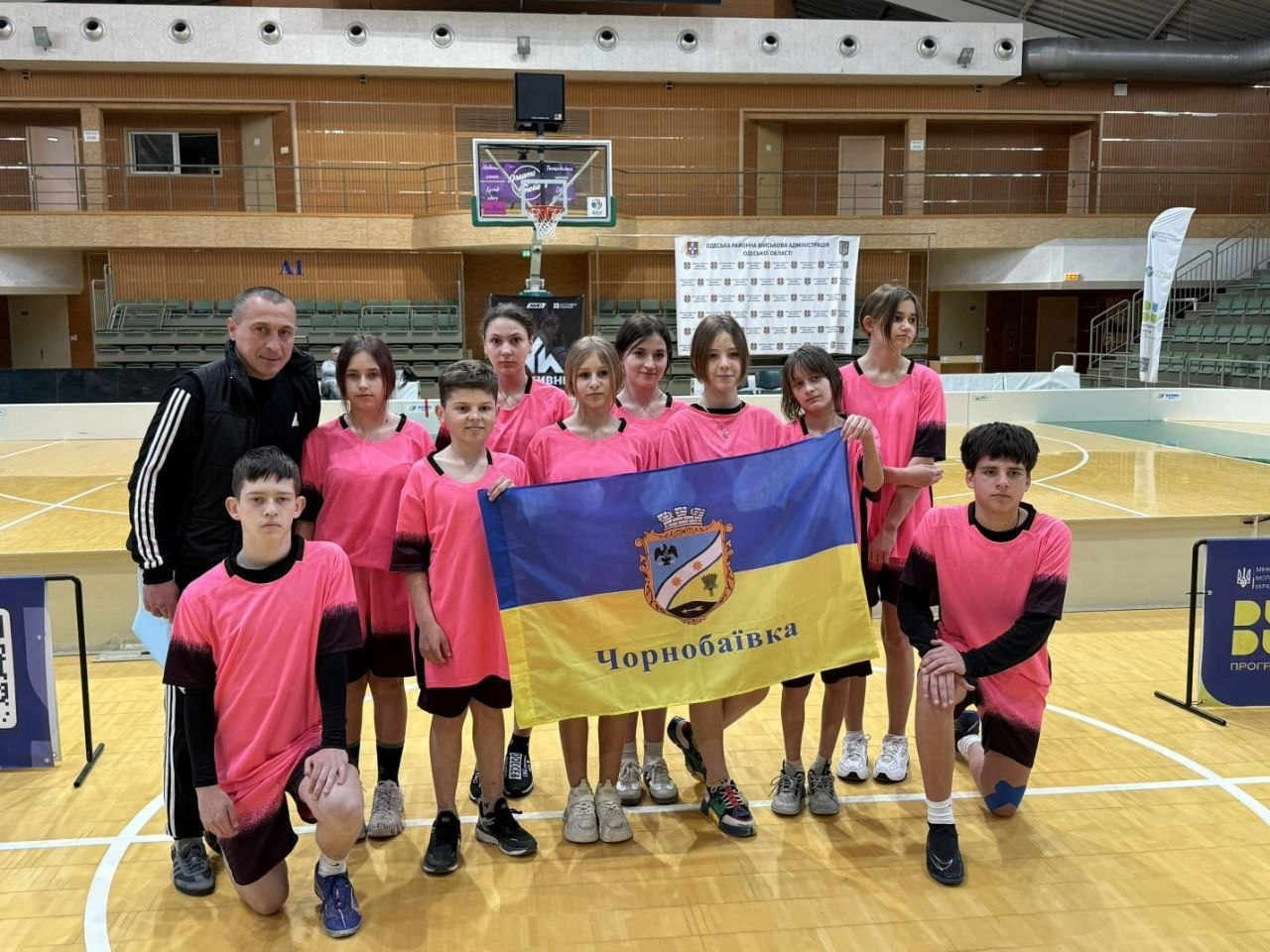Дитяча спортивна команда в рожевій формі разом із тренером тримають прапор Чорнобаївки на баскетбольному майданчику в спортзалі.