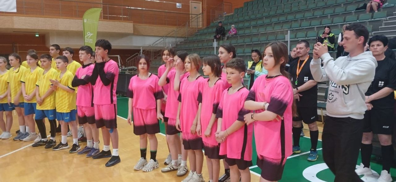 Дитячі спортивні команди в рожевій та жовтій формі вишикувалися на майданчику в спортзалі під час урочистої частини змагань. На задньому плані видно трибуни з глядачами.