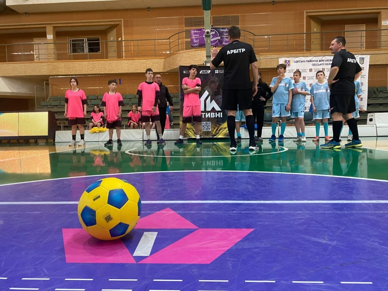 На передньому плані — жовто-синій м'яч у центрі спортивного майданчика. На фоні вишикувалися дві дитячі команди у рожевій та блакитній формі, а перед ними стоять арбітри в чорній формі.