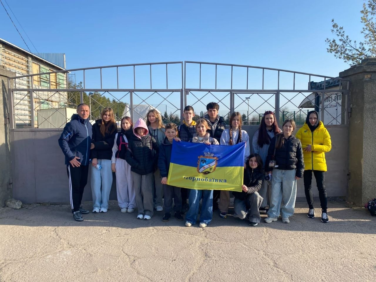 Група молодих людей разом із дорослим чоловіком позують для фото на фоні великих металевих воріт. У центрі вони тримають розгорнутий синьо-жовтий прапор Чорнобаївки.