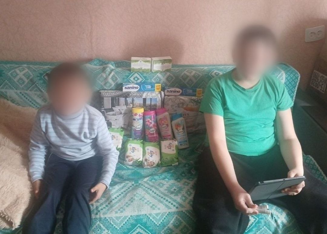 Двоє дітей із розмитими обличчями сидять на дивані поруч із набором засобів гігієни та побутових товарів, серед яких пакування підгузків, вологі серветки, шампуні та зубні пасти. Хлопчик праворуч тримає в руках планшет.