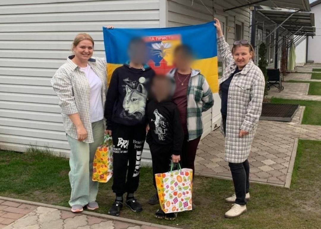 Три жінки та двоє дітей стоять біля будинку з розгорнутим прапором. У жінки, що стоїть поряд із дітьми, та в обох дітей обличчя розмиті. У руках дітей — яскраві подарункові пакети.