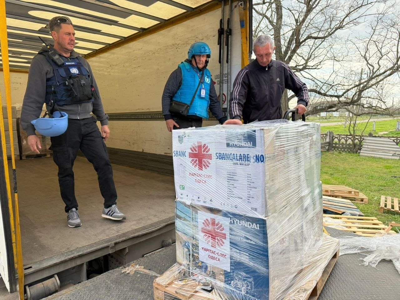 Троє чоловіків, двоє з яких у захисному спорядженні (бронежилетах та шоломах), розвантажують гуманітарну допомогу з вантажівки. На передньому плані — палета з коробками, на яких видно логотипи «Карітас» та Hyundai.