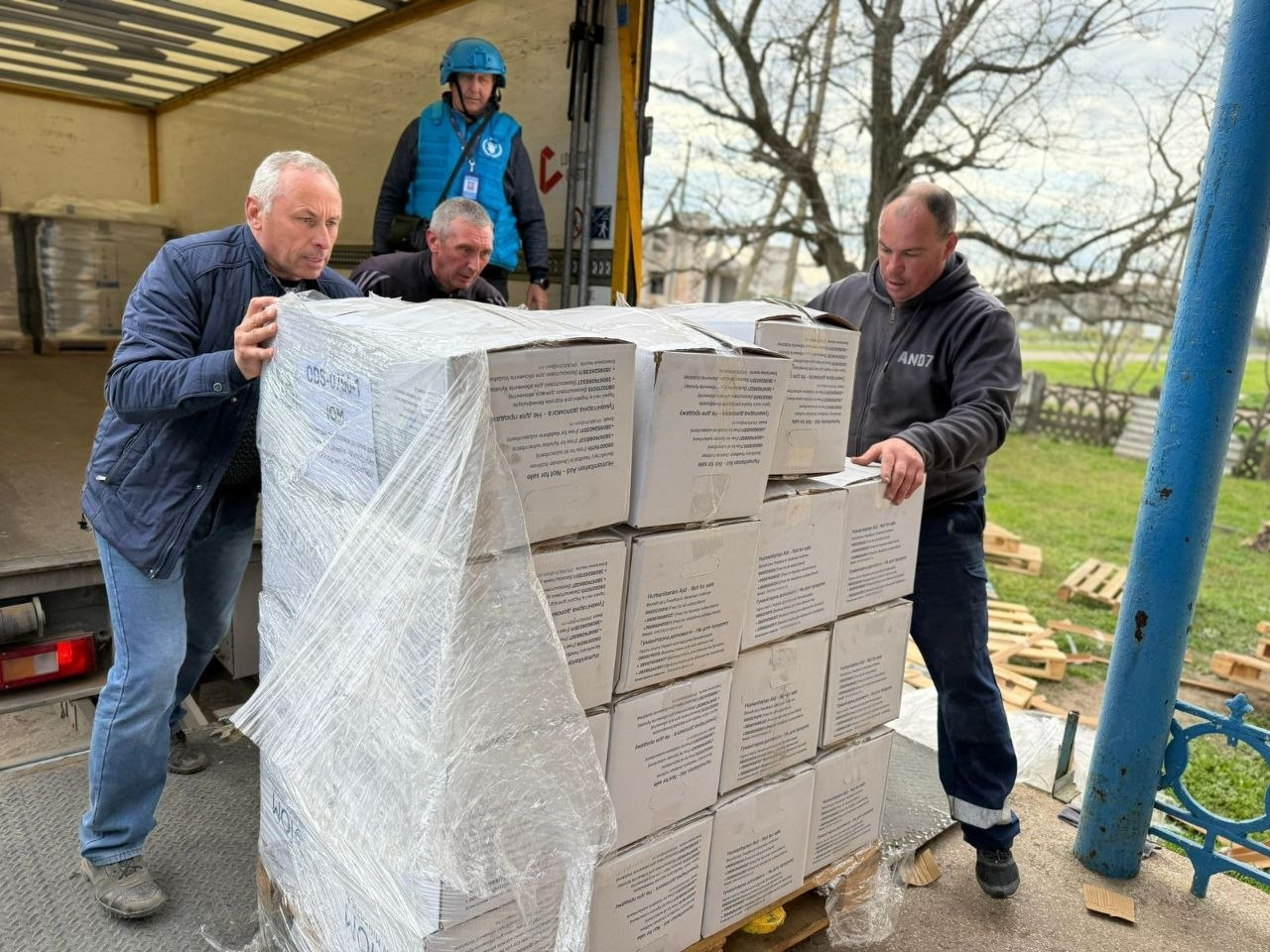 Четверо чоловіків розвантажують палету з коробками гуманітарної допомоги з кузова вантажівки. Один із присутніх одягнений у синій захисний жилет та шолом із символікою міжнародної організації.