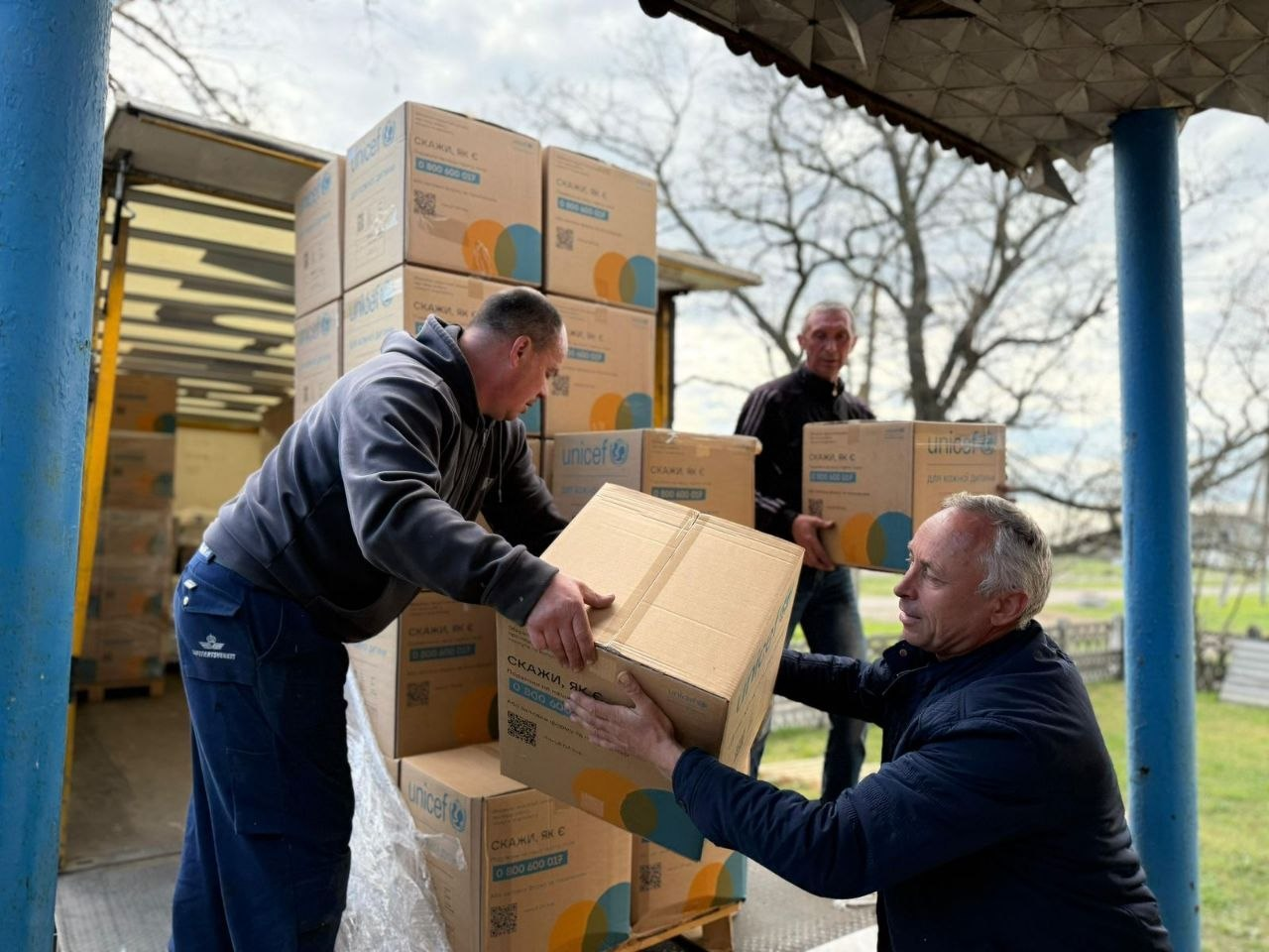 Троє чоловіків розвантажують картонні коробки з гуманітарною допомогою з логотипом UNICEF біля кузова вантажівки. Вони передають коробки один одному, складаючи їх у високу стопку на палеті.