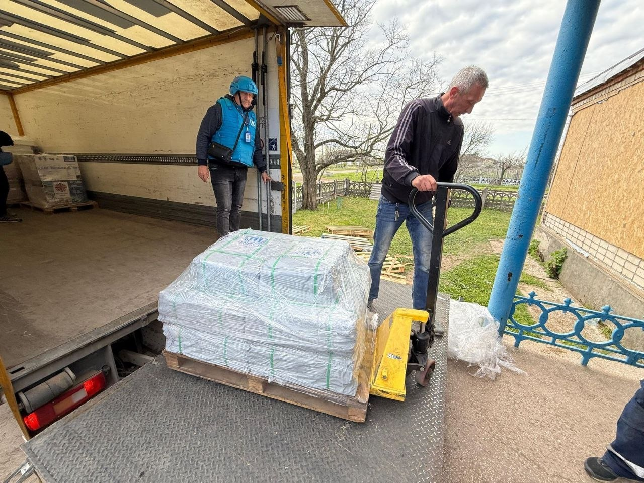Чоловік за допомогою гідравлічного візка (рокли) знімає з борту вантажівки палету з вантажем, обгорнутим плівкою. У кузові автомобіля стоїть інший чоловік у блакитному захисному жилеті та шоломі.