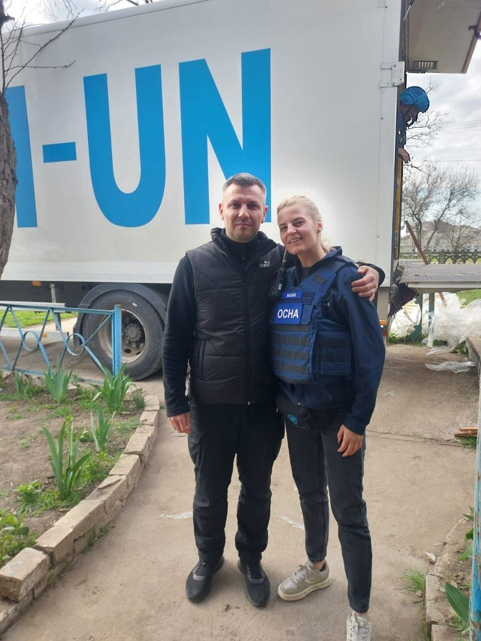 Чоловік та жінка в синьому захисному жилеті з написом «OCHA» позують на фоні білої вантажівки з великим (неповним) написом «ООН-UN». На задньому плані з кузова вантажівки визирає людина в захисному шоломі.