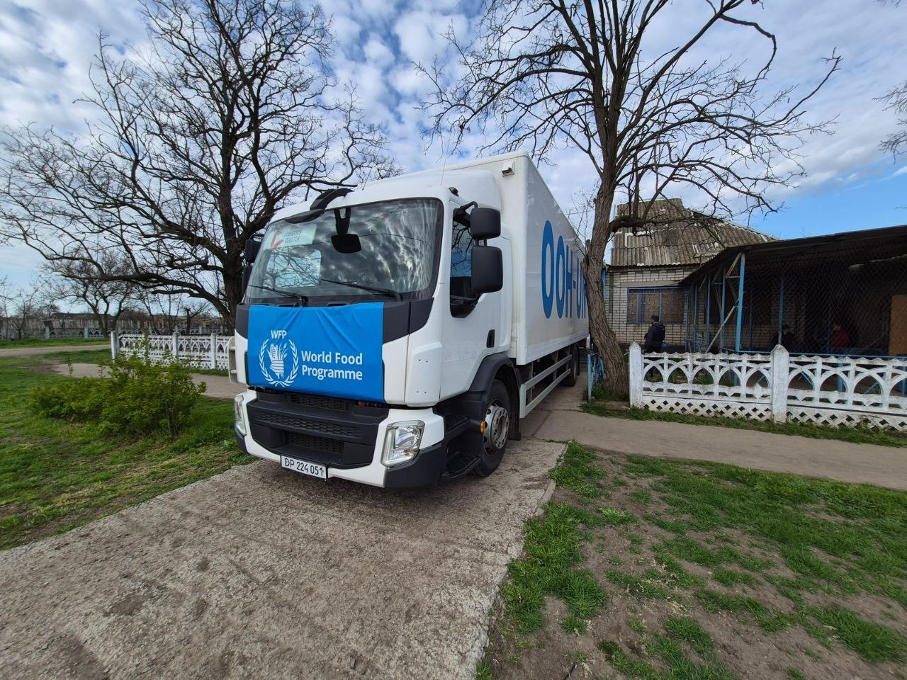 Біла вантажівка з синім банером «World Food Programme» на капоті та великим (неповним) написом «ООН-UN» на борту припаркована біля житлової забудови на сільській місцевості.