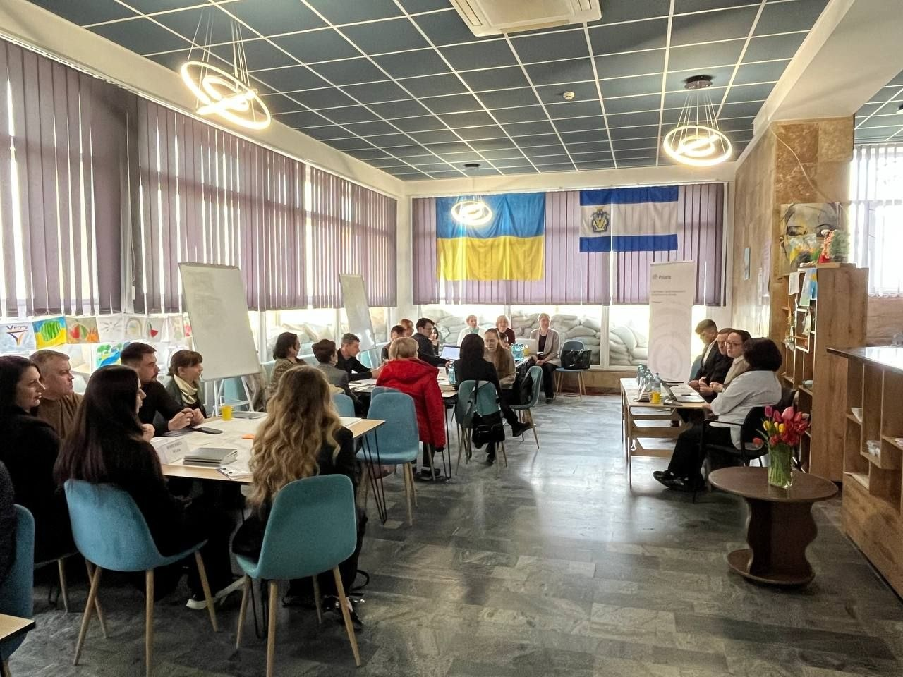 Група людей під час робочої зустрічі або тренінгу у великій залі. Учасники сидять за декількома столами, на стіні розміщено прапори, а вікна за ними захищені мішками з піском.