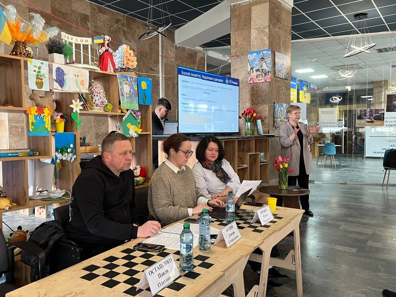 П’ятеро людей під час робочої зустрічі: троє сидять за столом-шахівницею, спікерка стоїть біля екрана з презентацією, а чоловік працює за комп’ютером на фоні.