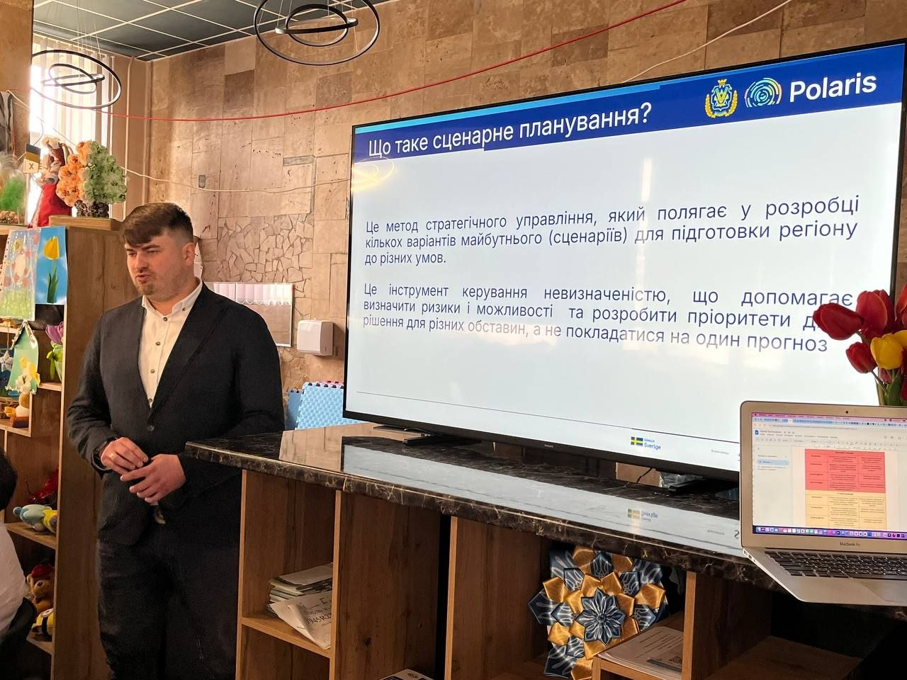Чоловік проводить презентацію на тему «Що таке сценарне планування?» у приміщенні з ноутбуком та великим екраном.