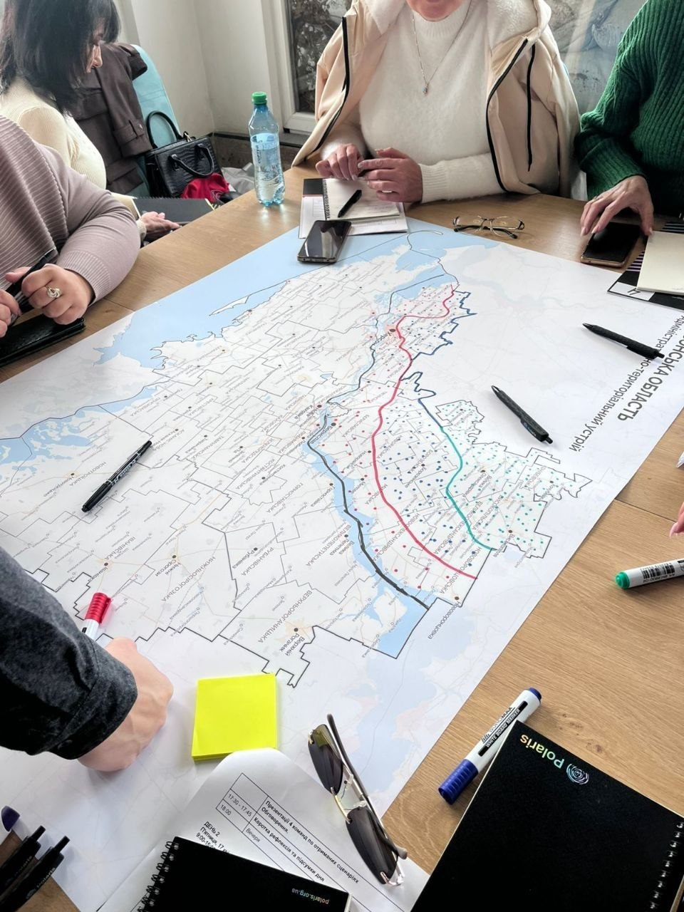 Група людей за столом працює з великою картою Херсонської області під час робочої наради. На столі розкладені маркери, блокноти та канцелярське приладдя.