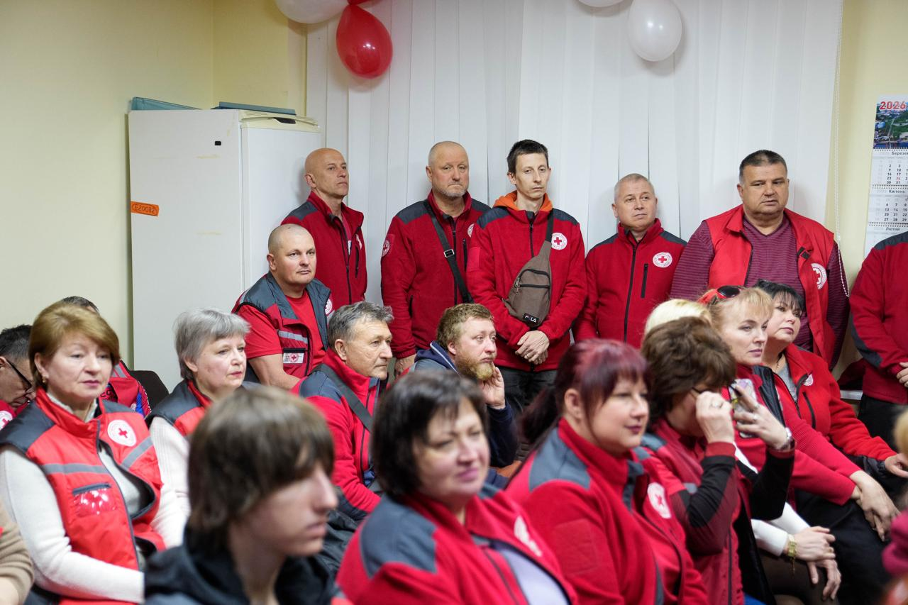 На фото група людей у приміщенні, більшість з яких одягнена в червоно-сірий формений одяг з емблемами Червоного Хреста. Деякі учасники сидять на передньому плані, інші стоять позаду на фоні білої стіни з червоними та білими повітряними кульками.