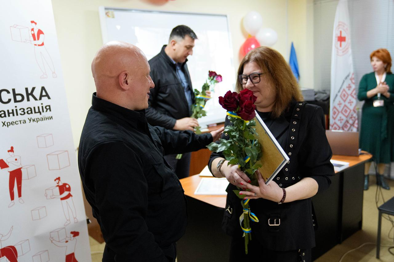 На фото чоловік у чорному одязі вручає жінці з каштановим волоссям в окулярах та чорній кофті букет червоних троянд і грамоту в рамці. Подія відбувається у приміщенні на фоні банера Товариства Червоного Хреста України та прапорів.