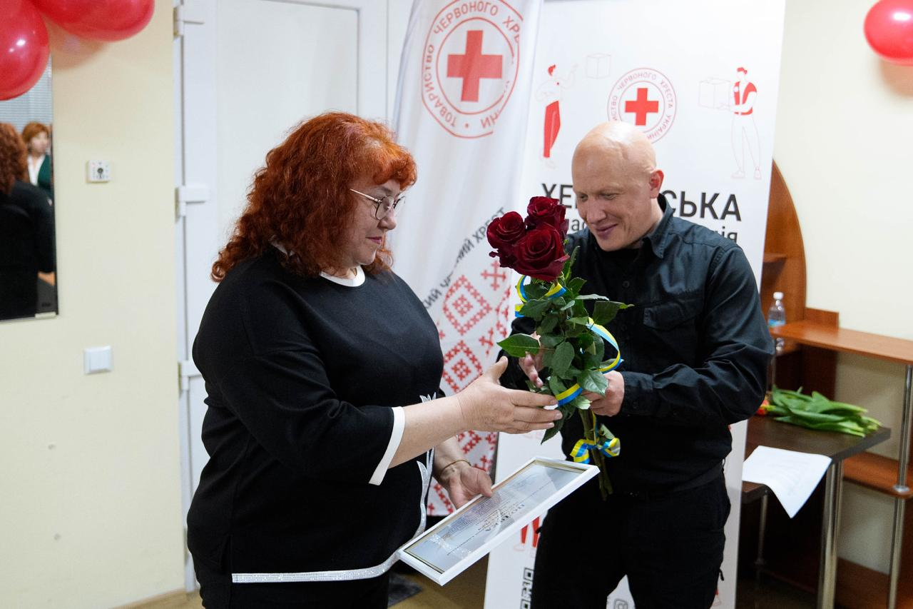 На світлині чоловік у чорному одязі вручає букет червоних троянд жінці з рудим волоссям в окулярах та чорній кофті. Жінка тримає в руках грамоту в рамці. Подія відбувається у приміщенні на фоні банера з логотипом Червоного Хреста.