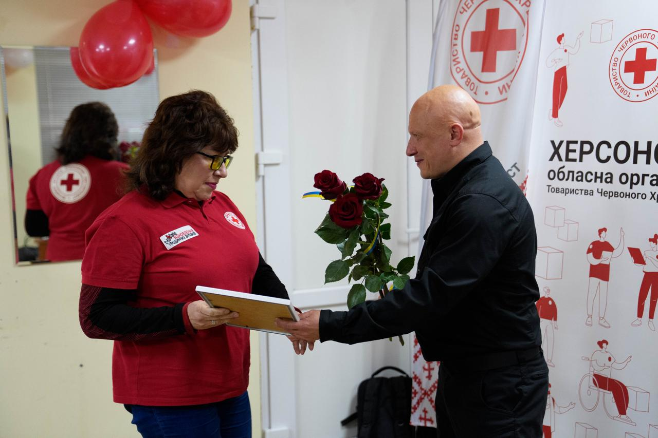 На фото чоловік у чорному одязі вручає грамоту в рамці та букет червоних троянд темноволосій жінці в окулярах та червоній футболці з емблемою. Подія відбувається у приміщенні поруч із банером Херсонської обласної організації Товариства Червоного Хреста України.