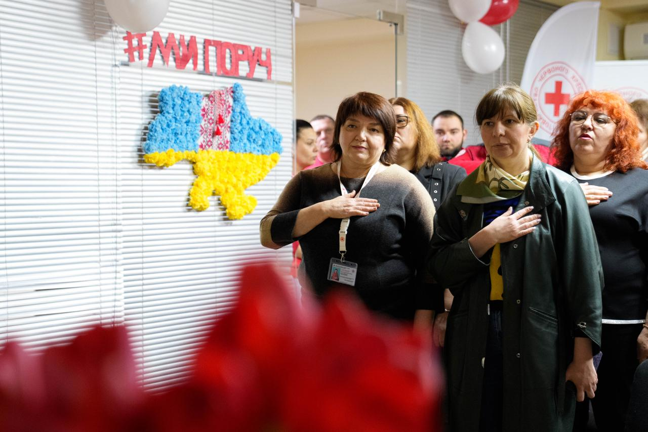 На фото група людей стоїть у приміщенні, приклавши праву руку до серця. На передньому плані — дві жінки: одна в темній кофті з бейджем, інша в зеленому плащі та жовто-блакитному шарфі. На задньому плані зліва видно мапу України у національних кольорах із написом «#МИПОРУЧ», а справа — прапор із логотипом Червоного Хреста.