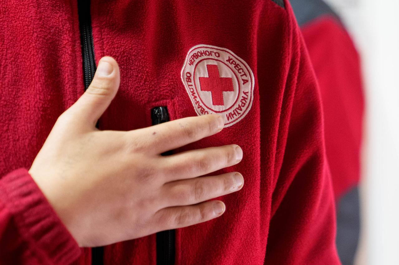 Крупний план руки, прикладеної до серця поверх червоної флісової куртки з емблемою Товариства Червоного Хреста України.