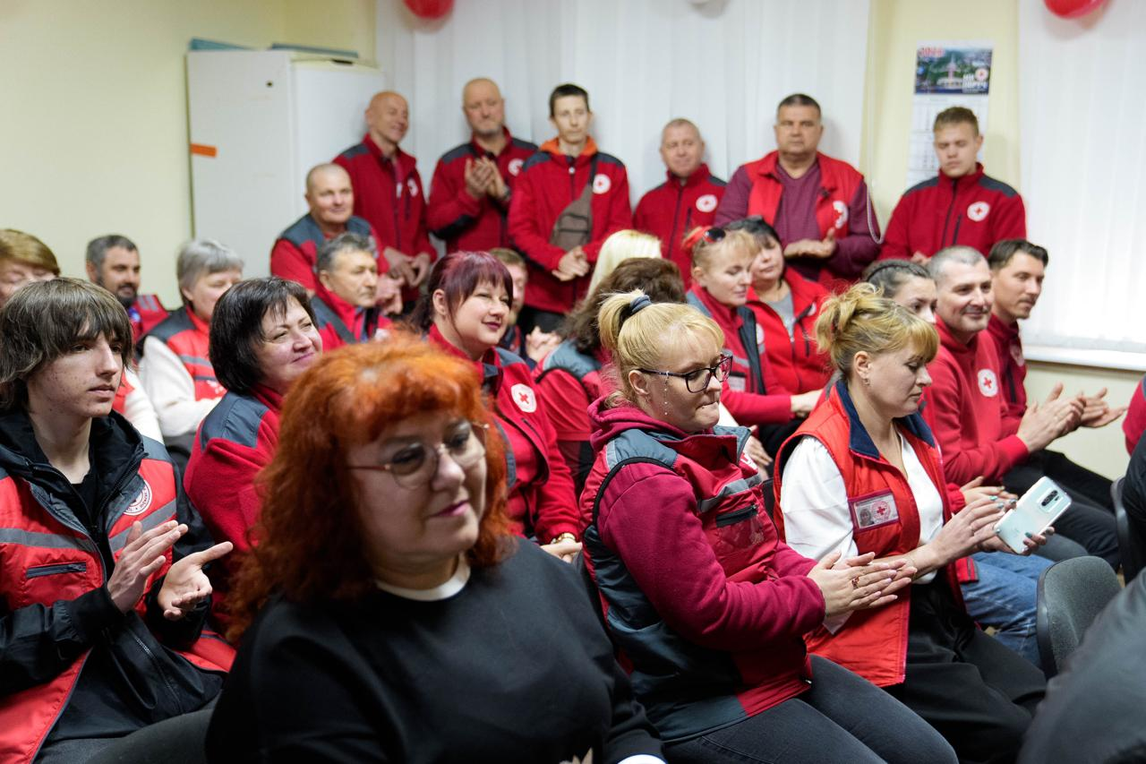 На фото група людей у приміщенні, більшість з яких одягнені в червоно-сірий формений одяг з емблемами Червоного Хреста. Люди сидять та стоять, дехто з них усміхається або аплодує. На передньому плані видно жінку з яскравим рудим волоссям в окулярах та чорній кофті.