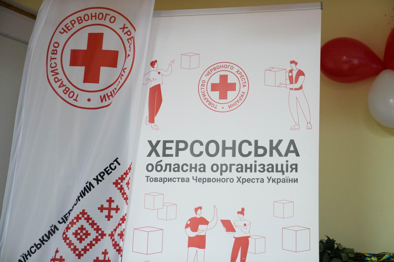 На фото зображено білий банер Херсонської обласної організації Товариства Червоного Хреста України з ілюстраціями волонтерів та коробок. Поруч стоїть білий прапор із логотипом організації та червоним традиційним орнаментом. На задньому плані видно червону та білу повітряні кульки.