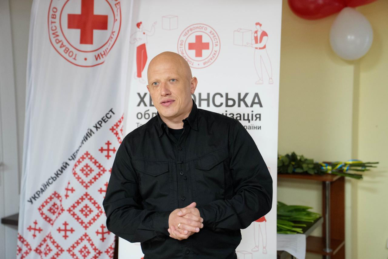 На фото чоловік у чорній сорочці стоїть у приміщенні на фоні банера Херсонської обласної організації Товариства Червоного Хреста України та прапора з логотипом організації. На задньому плані видно червону та білу повітряні кульки, а також квіти на столі.