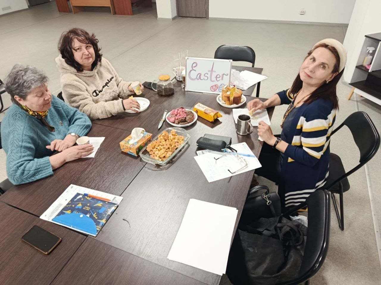 Три жінки сидять за столом у світлому приміщенні. На столі — частування, напої та табличка з написом «Easter». На передньому плані лежать папери, окуляри та смартфон.