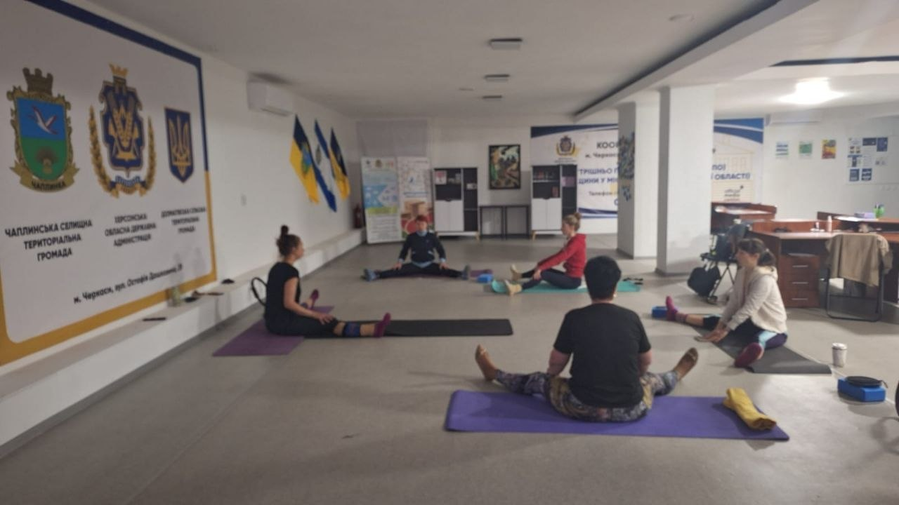 Група людей займається розтяжкою на килимках у просторому світлому залі. На стіні ліворуч зображені герби, а на задньому плані видно прапори України та інформаційні стенди.