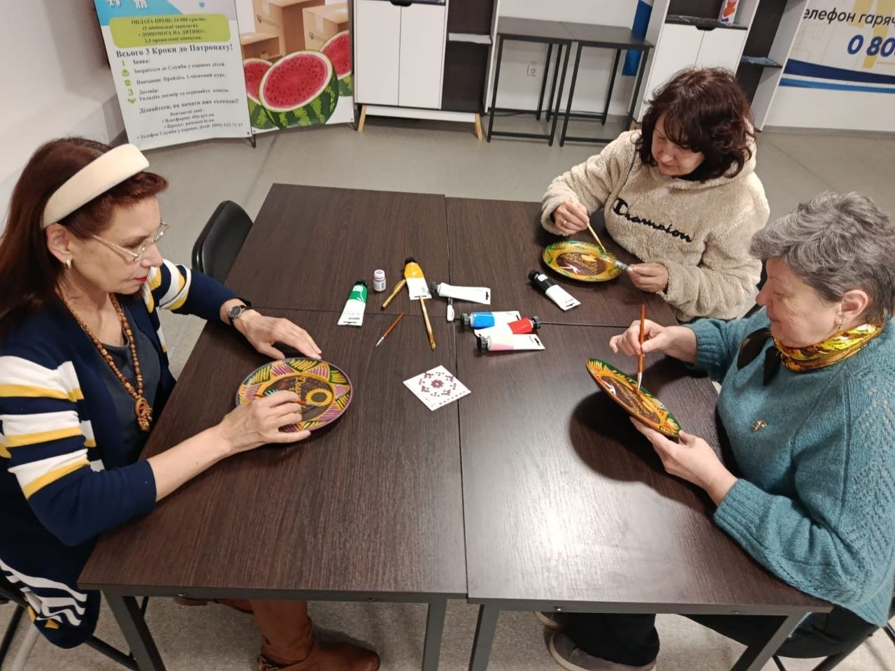 Три жінки за столом займаються розписом декоративних тарілок. На столі розкладені тюбики з фарбою, пензлі та зразок орнаменту. Подія відбувається у світлому приміщенні на фоні інформаційного стенда.
